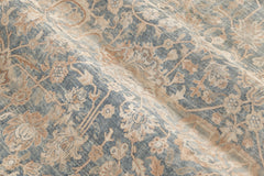 A close-up image of detailed textile, reminiscent of heirloom rugs. The Ben Soleimani KERMAN RUG, SOUTH EAST PERSIA, 10' 8" X 14' 8" features an intricate floral pattern with shades of beige and light brown over a muted blue background. The texture appears slightly crumpled, showcasing the ornate and delicate designs woven into the material.