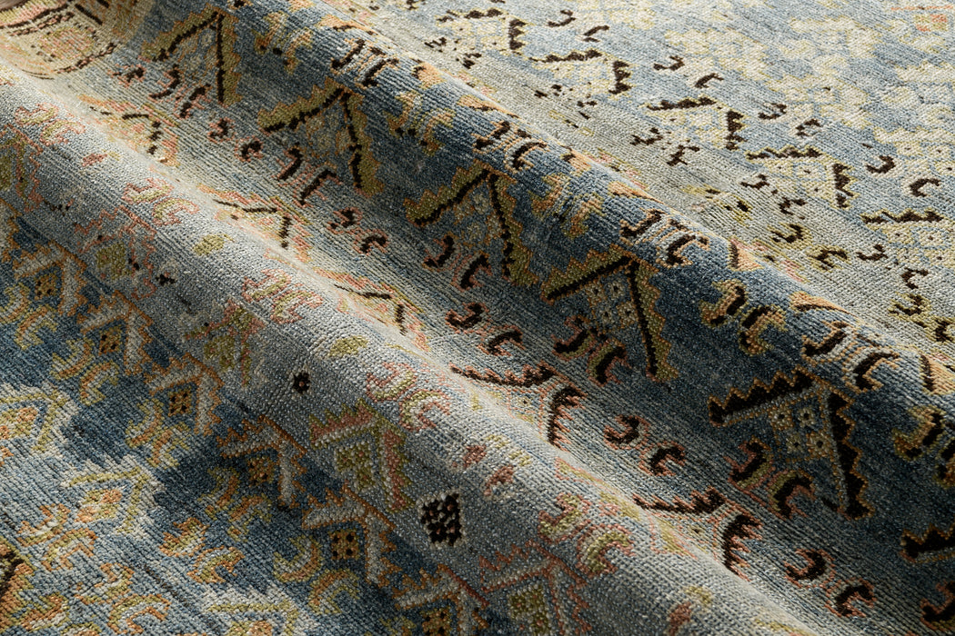 A close-up of an antique **MALAYIR RUG, AR30045/15857, WEST PERSIA, 4'4" X 6'9"** with intricate geometric and floral patterns. The heirloom piece by **Ben Soleimani** features a mix of colors including shades of blue, yellow, and brown. The fabric is slightly folded, showcasing the detailed design and texture.