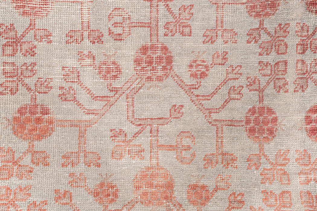 A close-up image of a woven textile featuring a symmetrical pattern of red floral and tree-like designs on a beige background. This heirloom-quality piece showcases intricate, vintage craftsmanship with repeated motifs spread across the fabric, reminiscent of antique rugs. The KHOTAN RUG, AR30065, EAST TURKESTAN, 5'11" X 11'1" by Ben Soleimani.