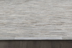 A close-up image of a beige and gray textured area rug from the Ben Soleimani collection features horizontal striations. The handcrafted Ladro Hide – Grey rug is placed on a dark wooden floor, with a small portion of its neatly finished edge visible in the bottom left corner.