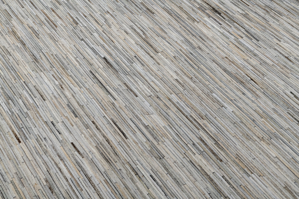 A close-up of a floor featuring a texture composed of narrow, elongated strips of various shades of gray, beige, and light brown, arranged in a linear pattern. The surface looks like it could be wooden or textile from the Ben Soleimani collection, providing a modern and minimalistic appearance. This is the Ladro Hide – Grey by Ben Soleimani.