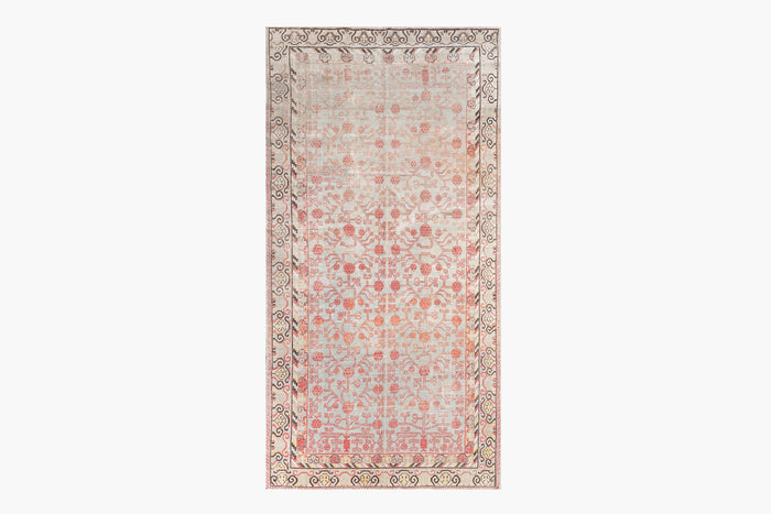 A rectangular rug featuring an intricate, symmetrical floral and geometric pattern in shades of light pink, beige, and taupe. The border has a repeating floral motif, and the central design includes stylized flowers and vines. With its distressed look, this heirloom-quality piece embodies the charm of antique rugs. This is the KHOTAN RUG, AR30065 from Ben Soleimani, measuring 5'11" x 11'1", hailing from East Turkestan.