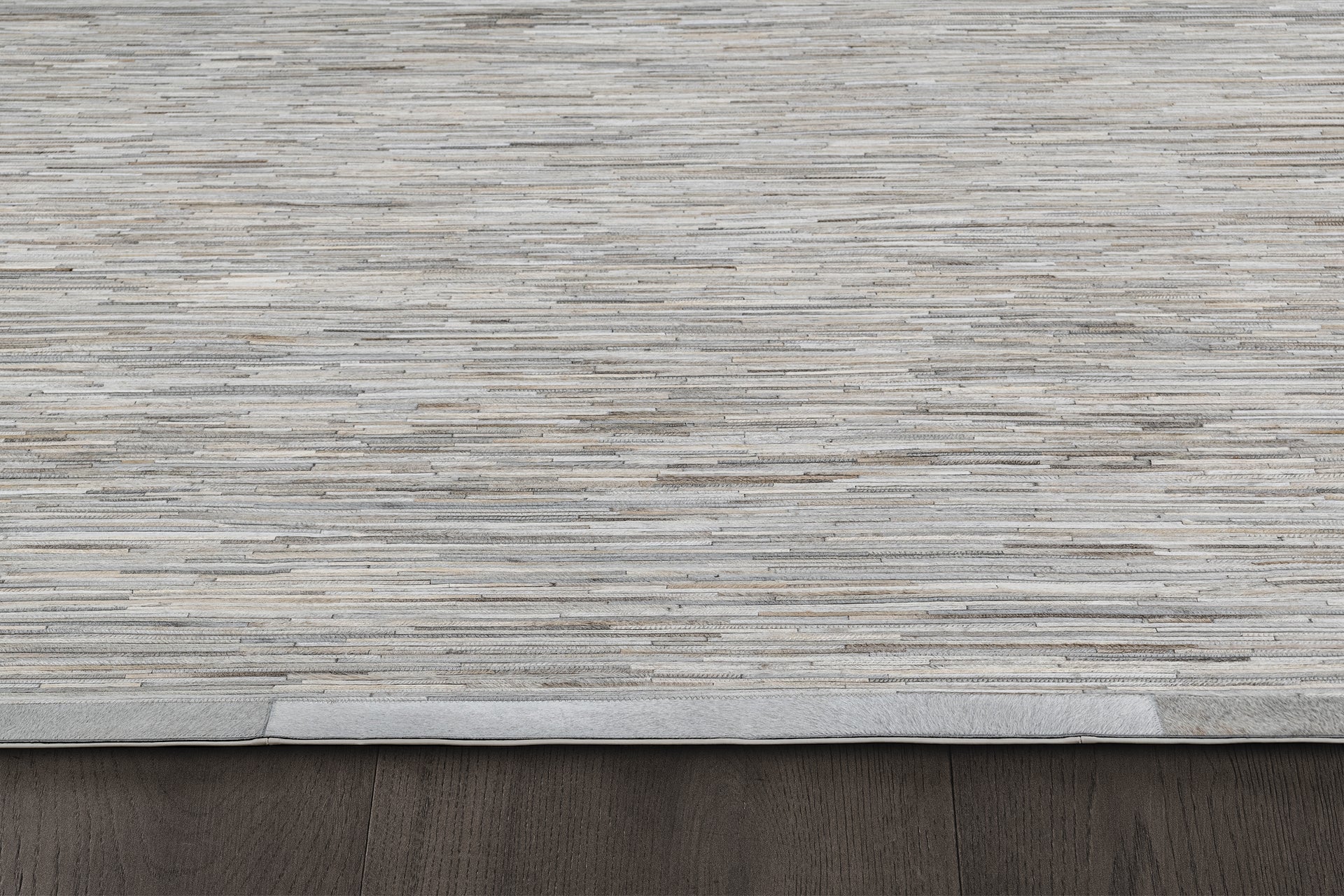 A close-up image of a beige and gray textured area rug from the Ben Soleimani collection features horizontal striations. The handcrafted Ladro Hide – Grey rug is placed on a dark wooden floor, with a small portion of its neatly finished edge visible in the bottom left corner.