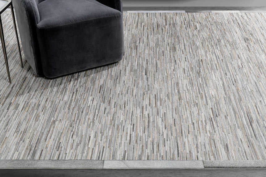 A gray velvet chair sits on a rectangular rug from the Ben Soleimani collection, showcasing narrow, multi-shade vertical stripes ranging from light to dark grays. The gray velvet chair and Ladro Hide – Grey by Ben Soleimani are positioned adjacent to a small metallic table on the left, against a smooth gray wall and floor.