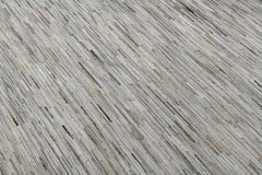 A close-up of a floor featuring a texture composed of narrow, elongated strips of various shades of gray, beige, and light brown, arranged in a linear pattern. The surface looks like it could be wooden or textile from the Ben Soleimani collection, providing a modern and minimalistic appearance. This is the Ladro Hide – Grey by Ben Soleimani.