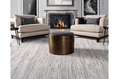 A modern living room features two beige armchairs with dark cushions facing each other, a dark cylindrical coffee table in between, and a lit fireplace with an ornate mantel in the background. The room has light grey walls, framed artwork above the fireplace, and a Ladro Hide – Grey from the Ben Soleimani collection.